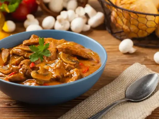 Aksamitny strogonow z pieczarkami w niebieskiej misce, udekorowany natką pietruszki. Obok leży łyżka na lnianej serwetce.