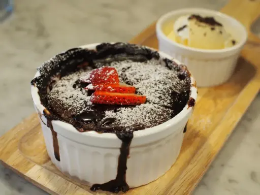 Pyszny, gorący lava cake z mikrofali, posypany cukrem pudrem i udekorowany truskawkami. Obok porcja lodów waniliowych.