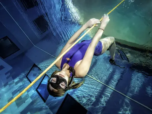 Kobieta w fioletowym stroju kąpielowym i masce nurkuje w basenie, trzymając się liny. To przykład tego, freediving co to jest – sport wymagający kontroli oddechu i siły.