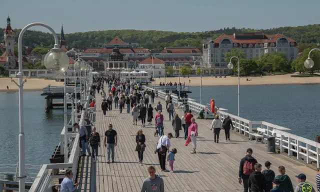 Tłum ludzi spaceruje po molo w Sopocie, jednym z nietypowych atrakcji Gdańska. W tle widać plażę i zabytkowe budynki.