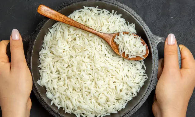 Dwie ręce trzymają czarną patelnię z puszystym ryżem. Drewniana łyżka pokazuje, jak ugotować ryż na parze.