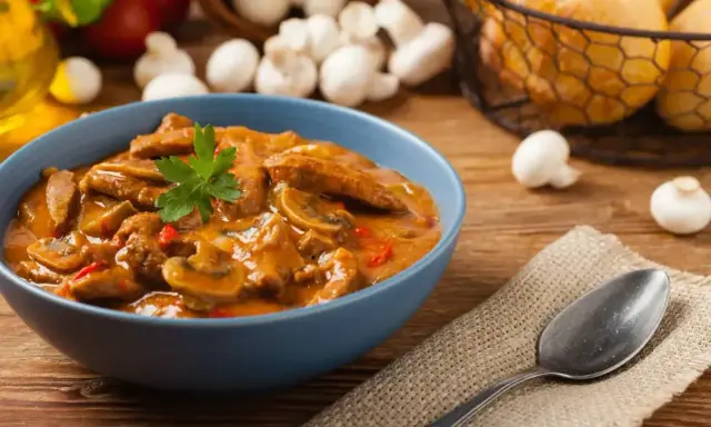 Aksamitny strogonow z pieczarkami w niebieskiej misce, udekorowany natką pietruszki. Obok leży łyżka na lnianej serwetce.